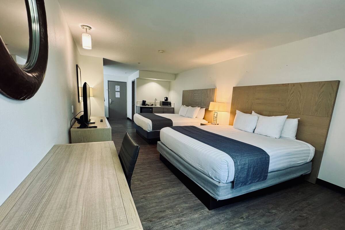 A hotel room with two beds, a desk, a chair, a mirror, a small kitchenette, and a bathroom.