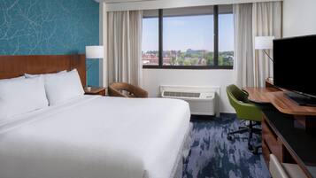 A hotel room with a large bed, a desk with a flat-screen TV, a chair, a desk lamp, and a view of the city through the window.
