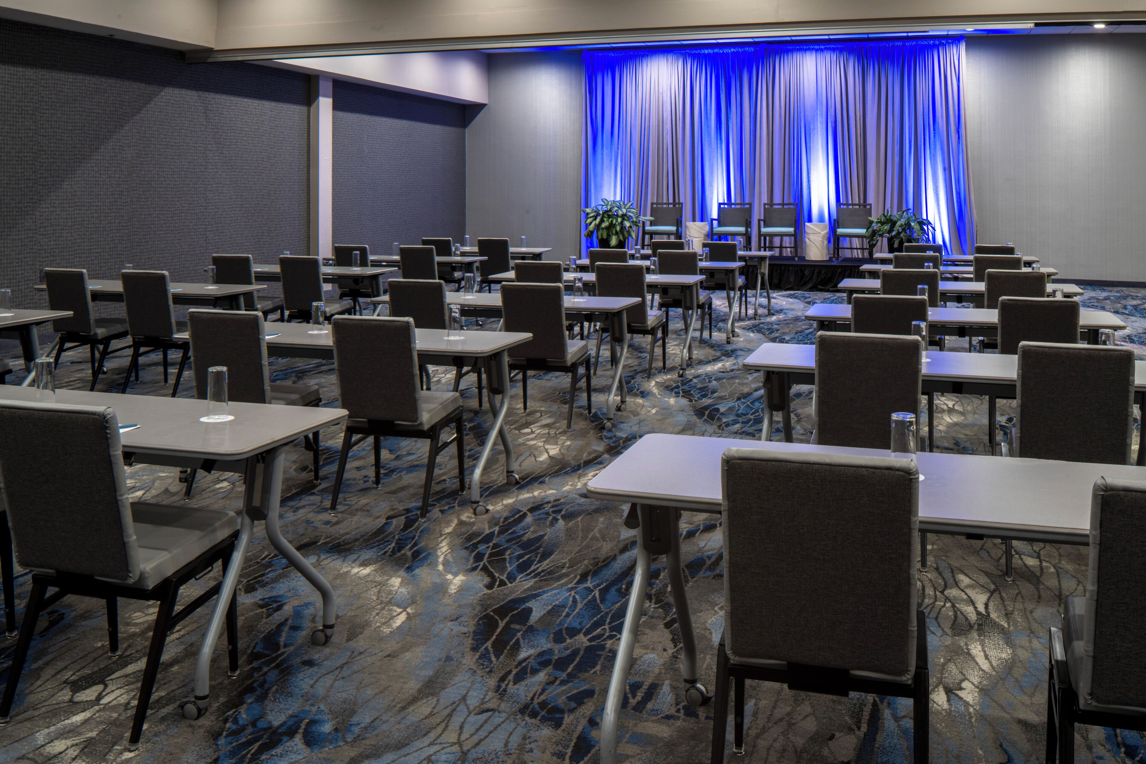 A conference room with rows of tables and chairs, a stage with a blue curtain backdrop, and a carpeted floor.