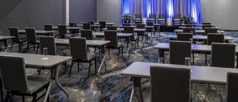 A conference room with rows of tables and chairs, a stage with a blue curtain backdrop, and a carpeted floor.