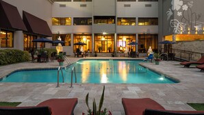 A swimming pool with lounge chairs and umbrellas, surrounded by a building with multiple floors and large windows.