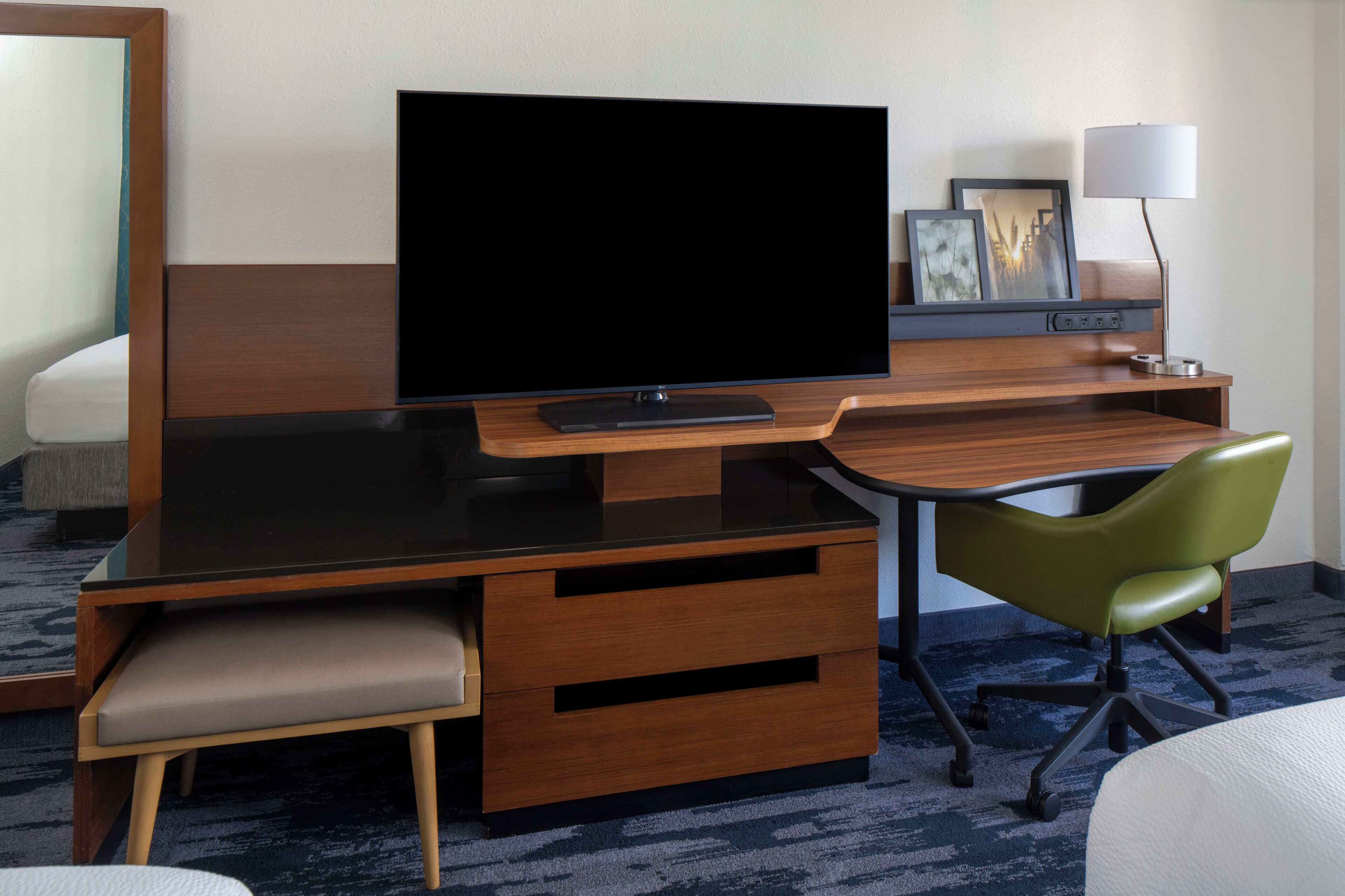 Ein Hotelzimmer mit Flachbildfernseher, Schreibtisch mit Stuhl, Nachttisch, Lampe und Bett.