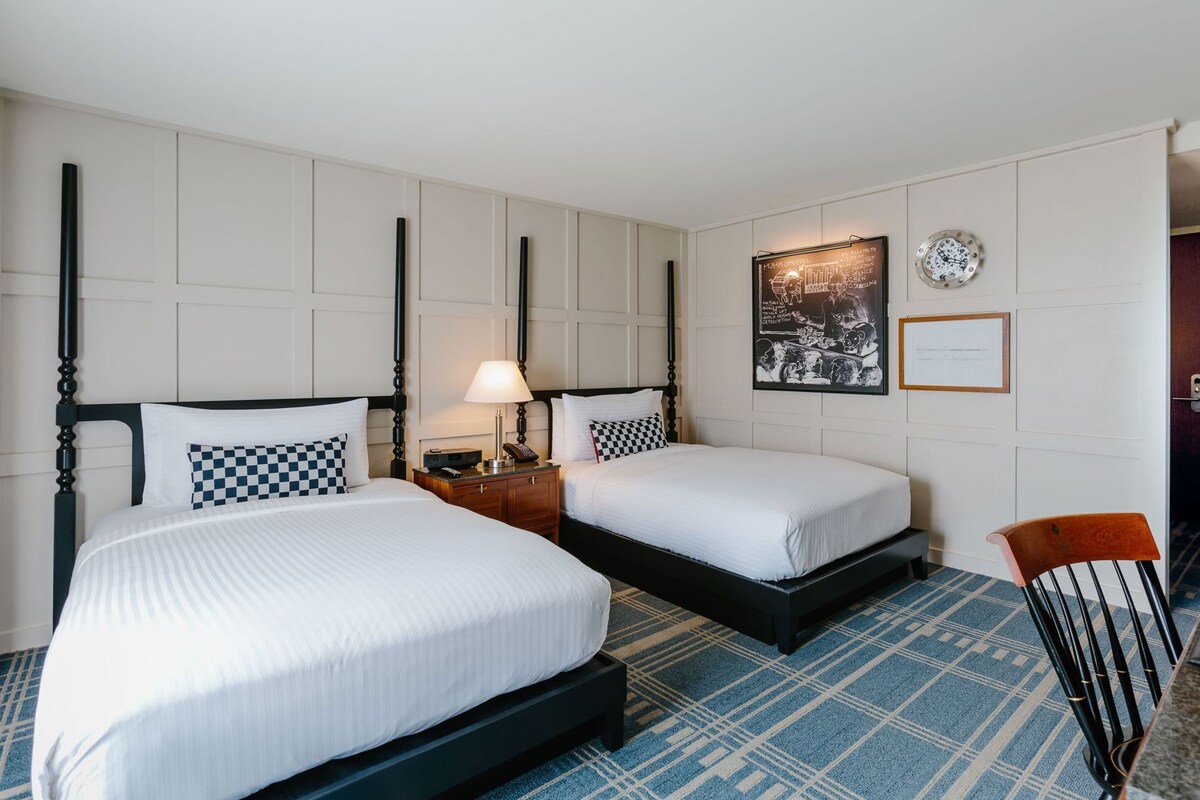 A hotel room with two beds, a nightstand, a lamp, a framed picture, a clock, and a chair.