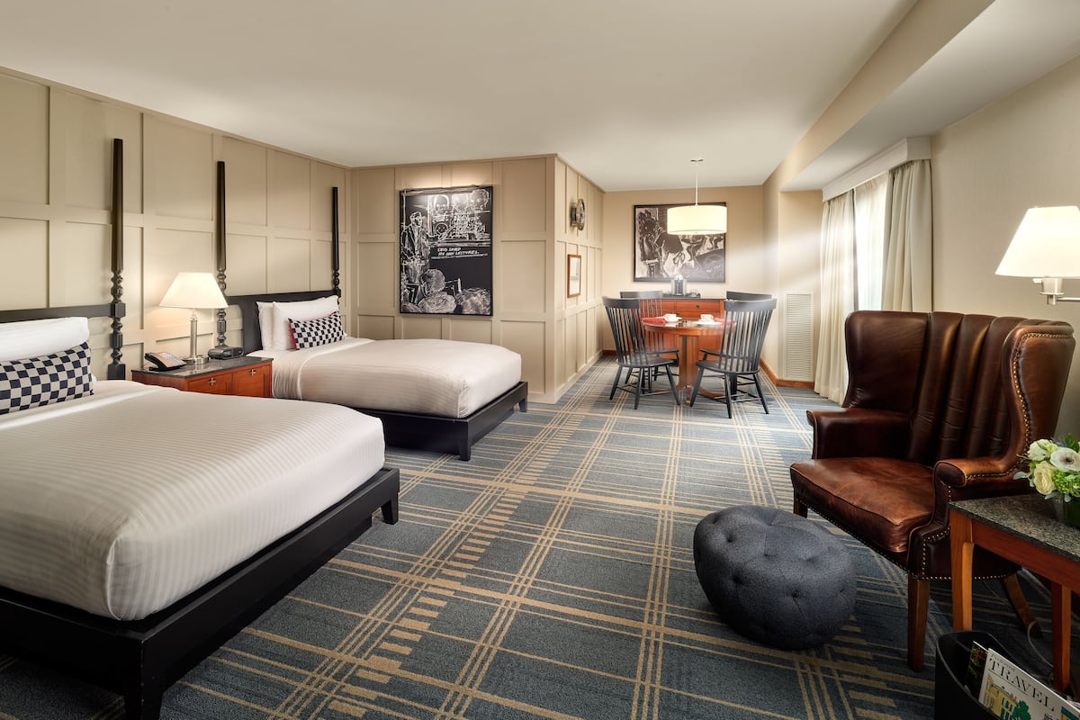 A hotel room with two beds, a desk with chairs, a leather armchair, and a small ottoman.