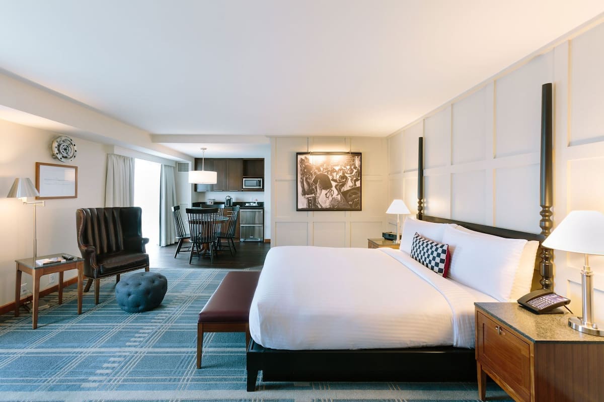 A hotel room with a large bed, a desk, a chair, a dining table, and a kitchen area.