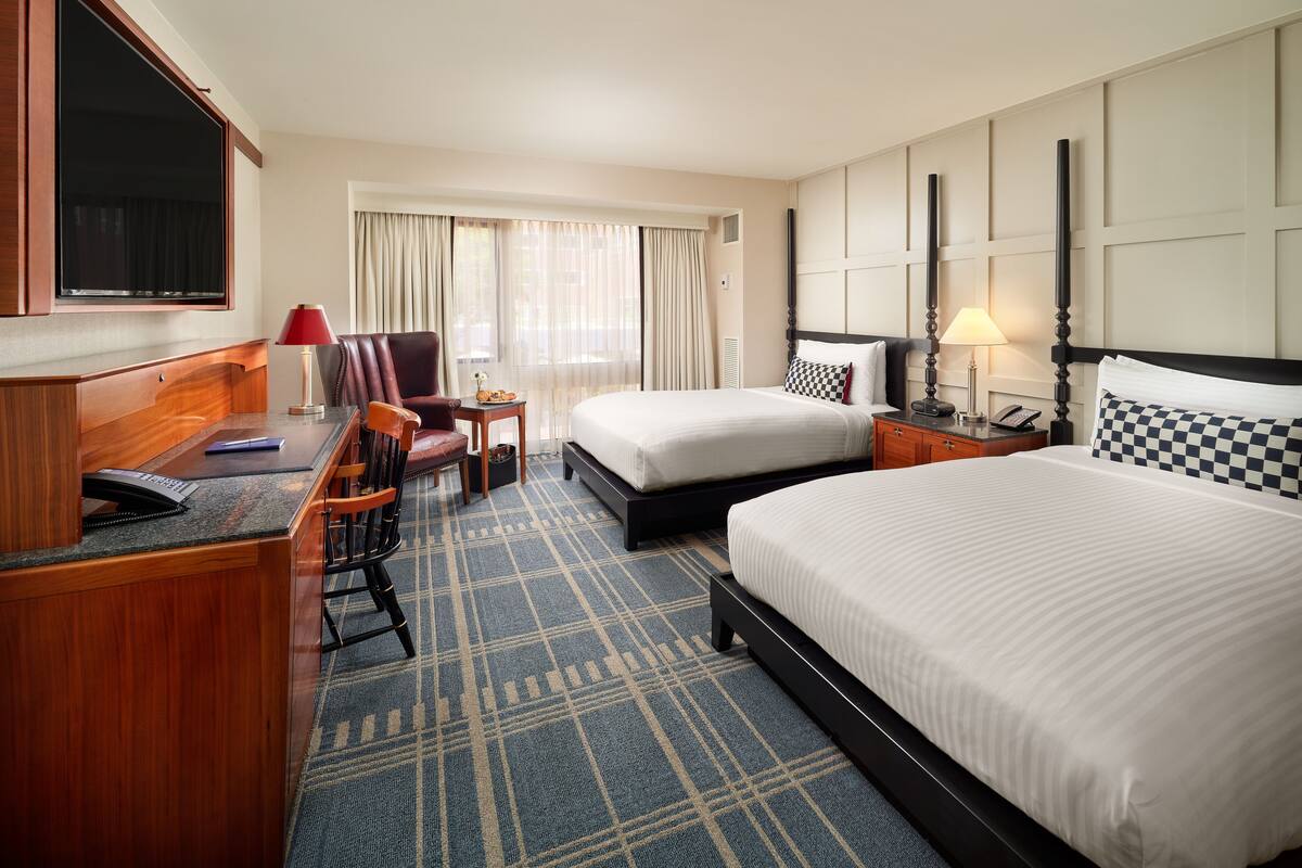 A hotel room with a desk, two beds, a television, and a window with curtains.