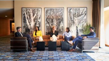 A group of five people seated on a brown sofa in a lobby with large abstract tree paintings on the wall, a patterned carpet, and a large window with curtains.