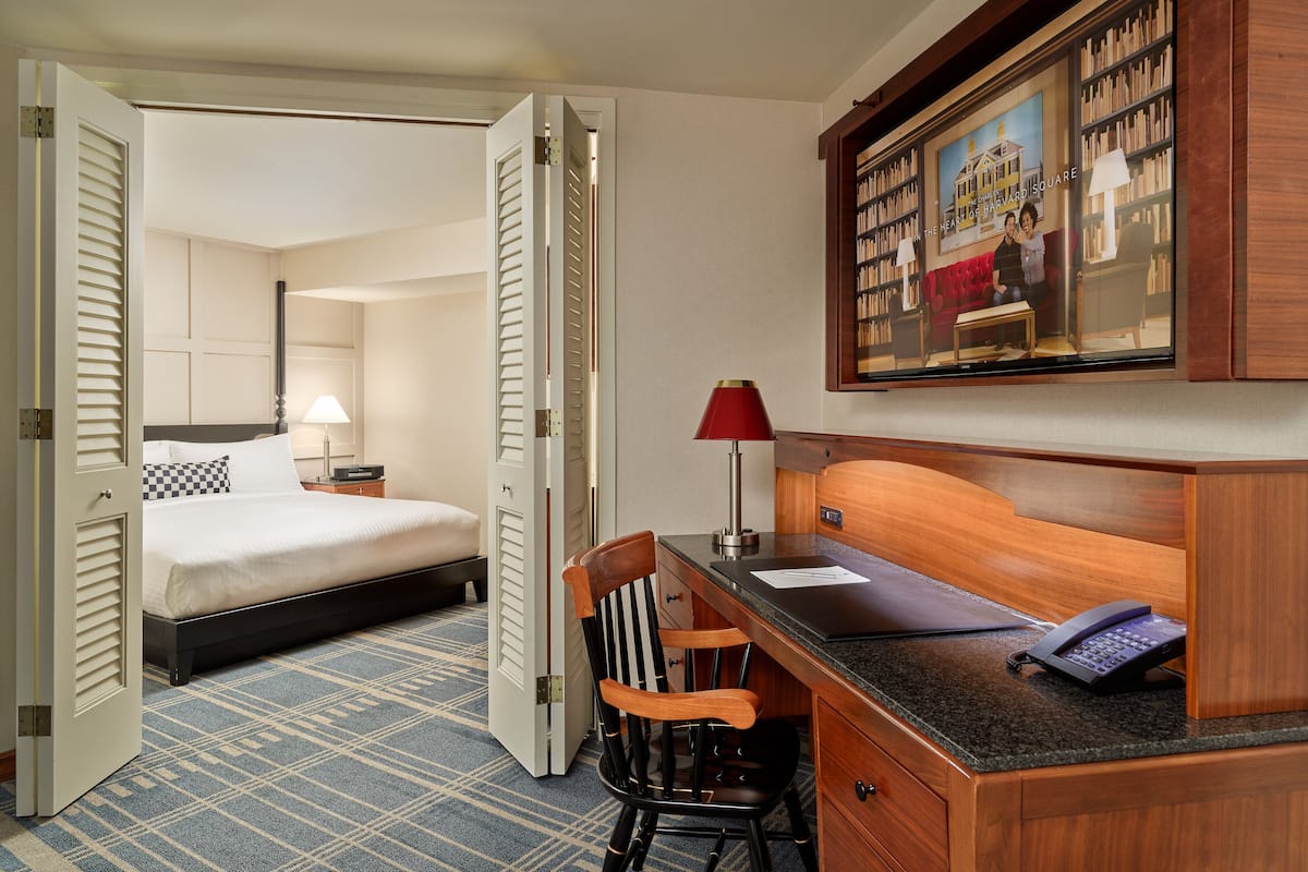 A hotel room with a bed, desk, chair, and a painting of a couple on the wall.