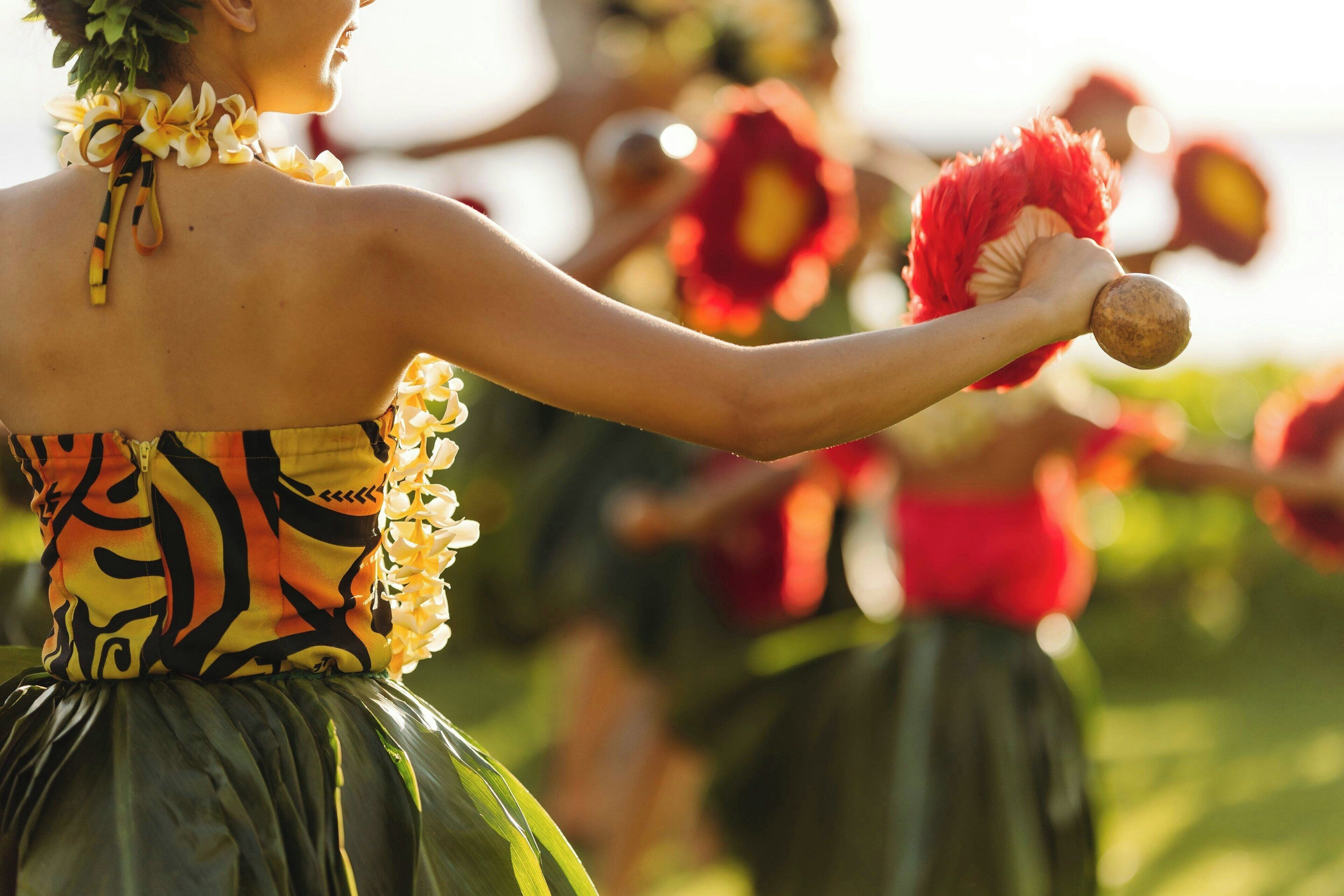 Une personne vêtue d’une tenue tropicale, portant un collier de fleurs et tenant un éventail rouge.