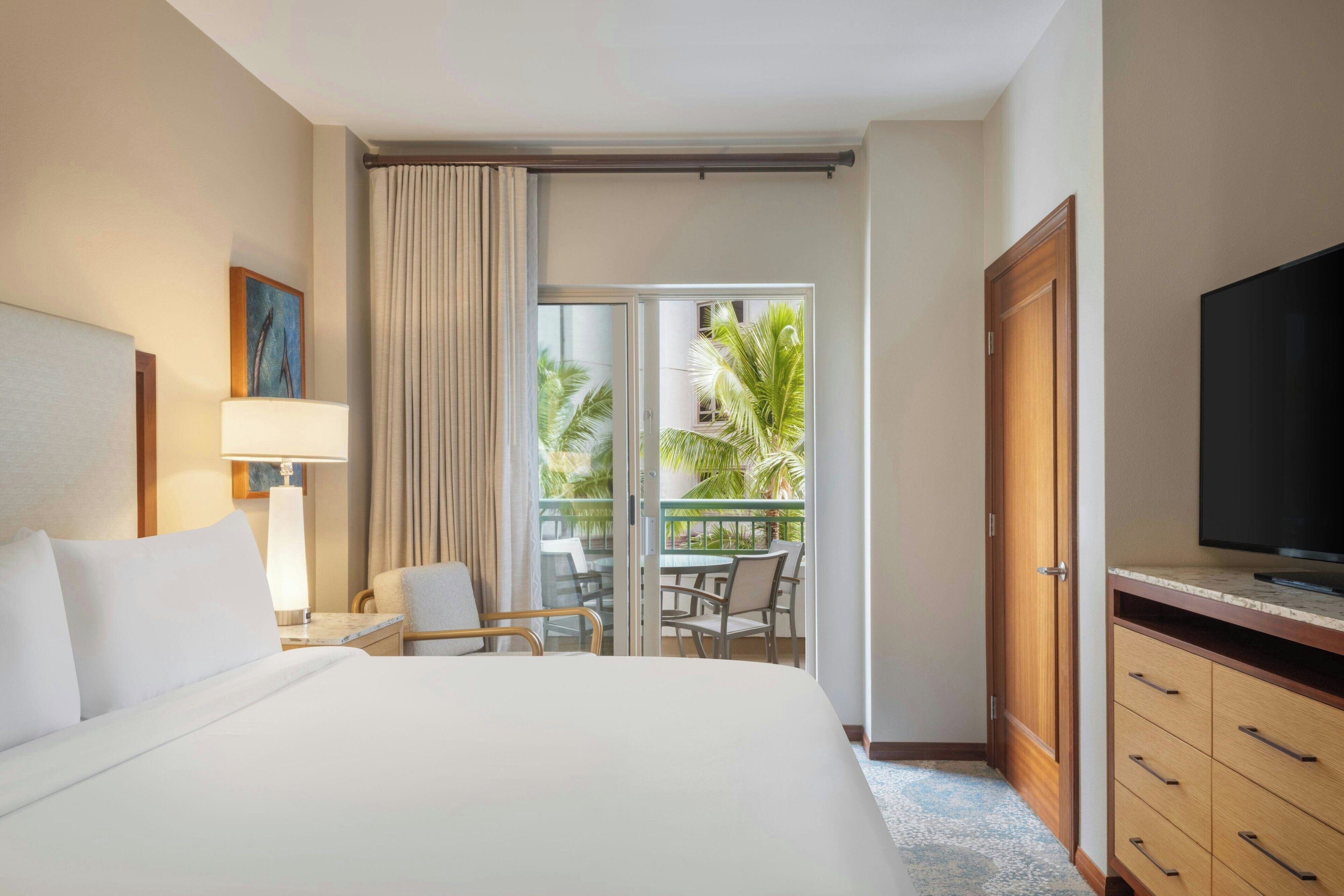 A hotel room with a bed, a TV, a desk with a lamp, a chair, a balcony with a view of palm trees, and a sliding door.
