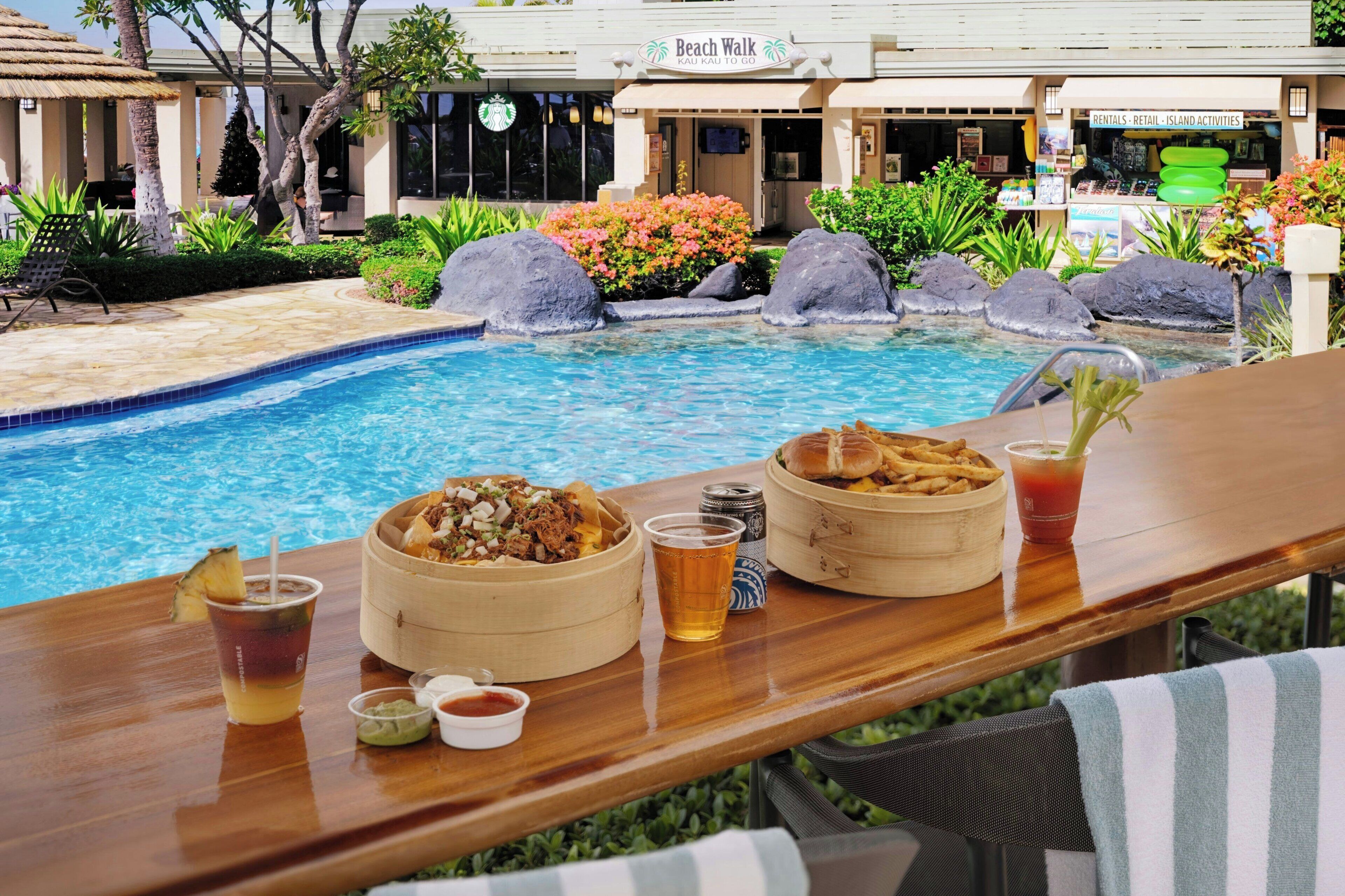Un bar junto a la piscina con comida y bebidas, un letrero que indica un paseo por la playa y una tienda de alquileres.