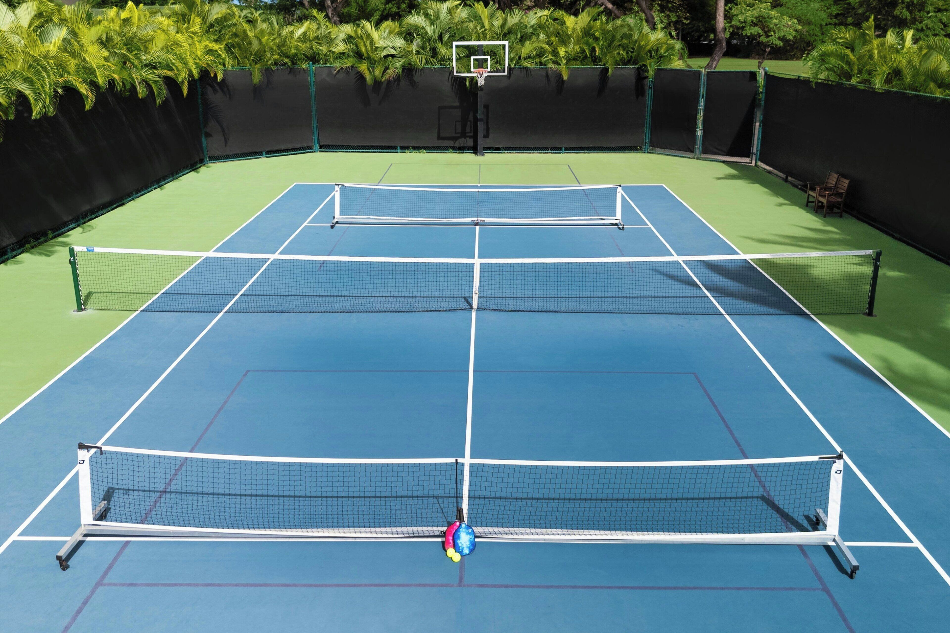 A tennis court with a net, surrounded by a green fence and palm trees.