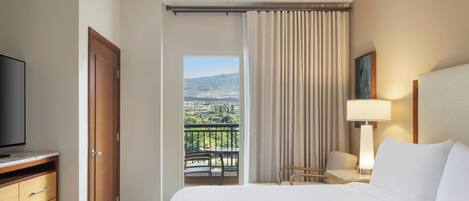A hotel room with a bed, a TV, a desk with a chair, a lamp, and a view of a city and mountains through a window.