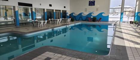 An indoor swimming pool with a wave design on the walls, a seating area with chairs, and a sign indicating pool hours.