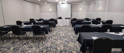 Un salón de conferencias con varias mesas y sillas, una pantalla de proyector y piso alfombrado.