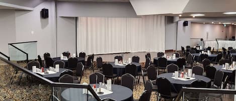 A conference room with multiple tables and chairs, a stage with a curtain, and a carpeted floor.