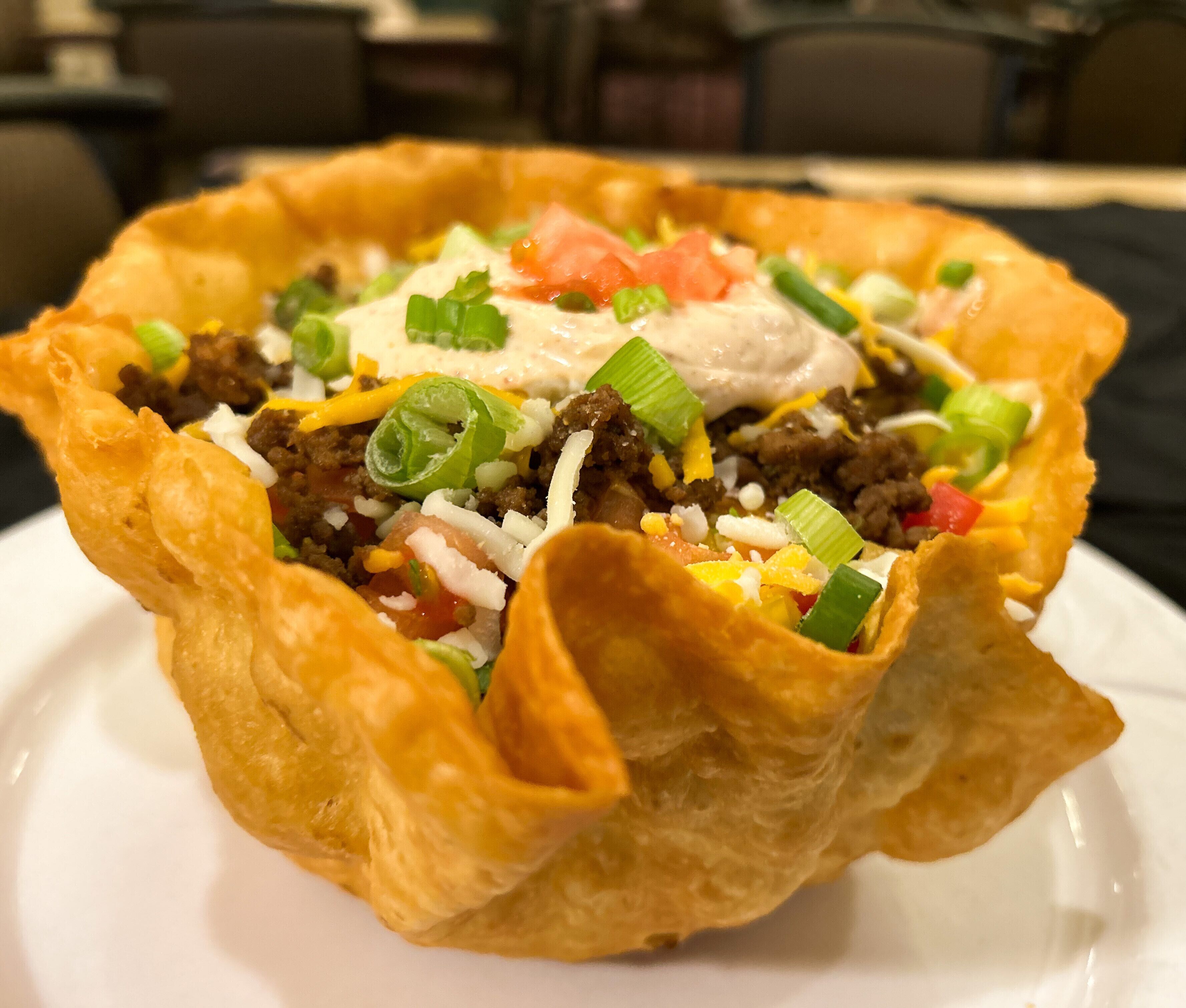 A taco bowl with ground beef, cheese, and toppings on a white plate.