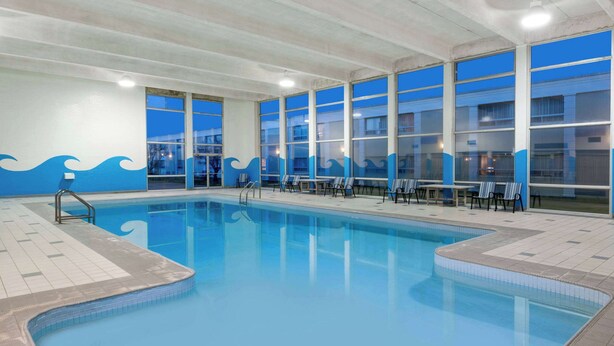 An indoor swimming pool with clear blue water, surrounded by lounge chairs and tables, and large windows with a view of the outside. - Ramada by Wyndham London (London)