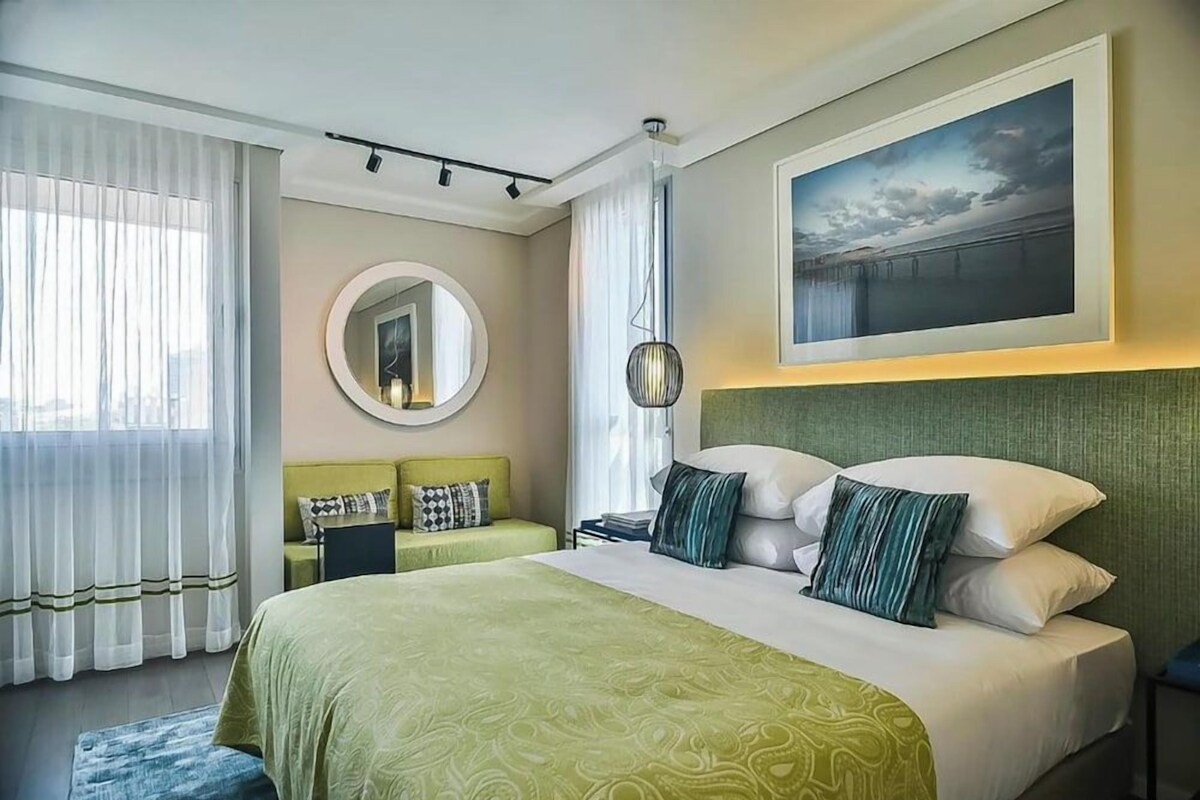 Hotelkamer met een groot bed, een ronde spiegel, een groen hoofdeinde, een schilderij en uitzicht op de stad.