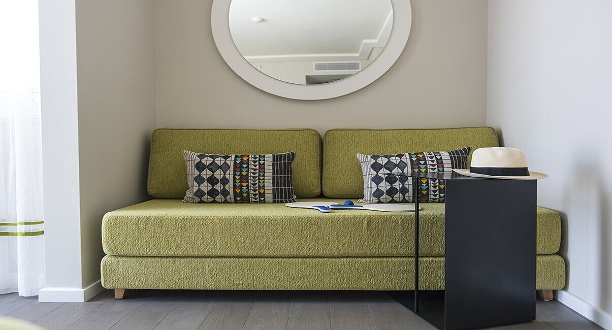 A green sofa with patterned pillows, a round mirror, and a black side table with a hat.