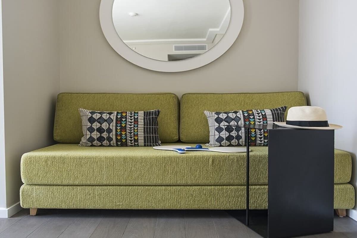 A green sofa with patterned pillows, a round mirror, and a black side table with a hat.