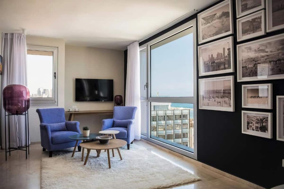 Ein modernes Wohnzimmer mit zwei blauen Sesseln, einem Couchtisch, einem Flachbildfernseher und einem großen Fenster mit Blick auf die Stadt.