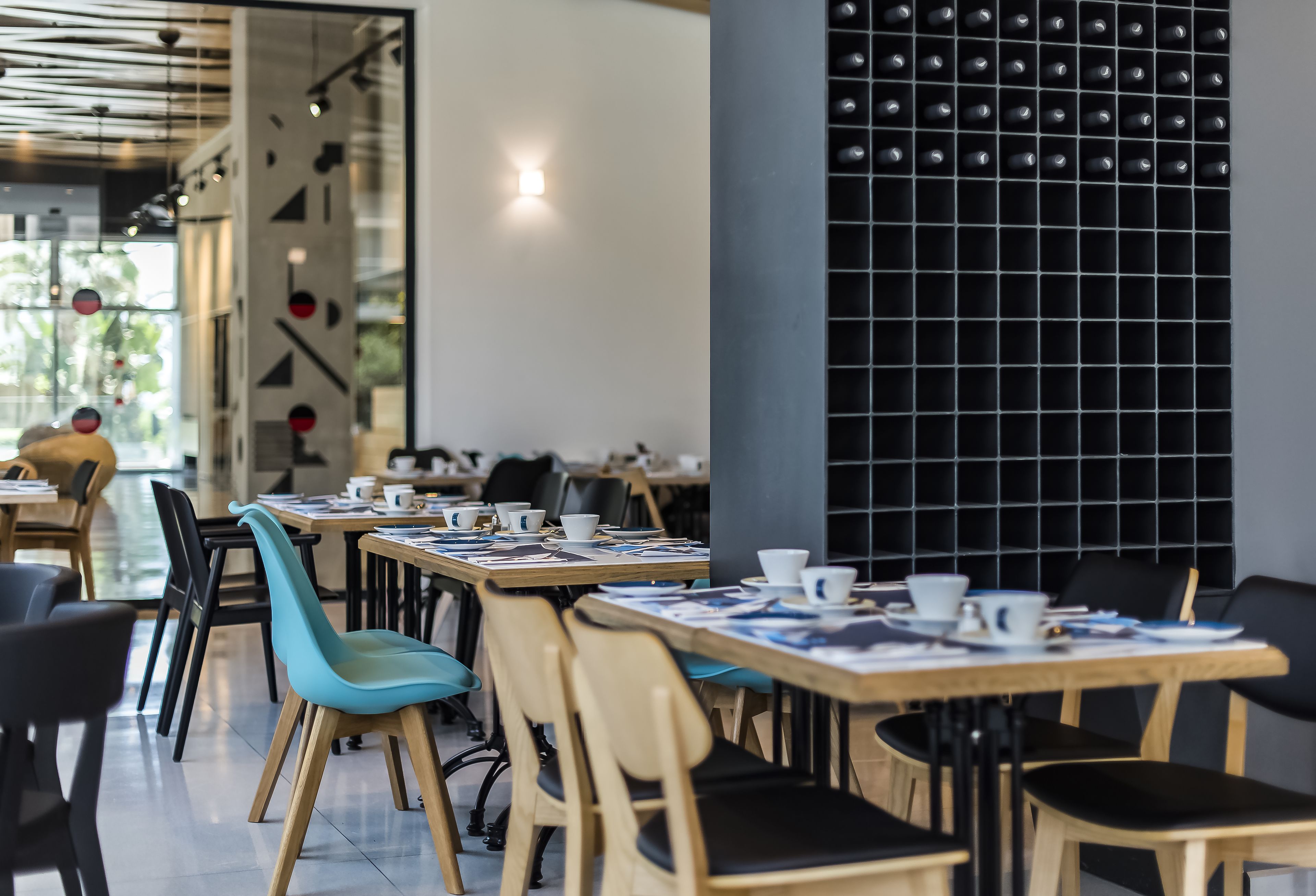 Un café moderne avec des tables et des chaises en bois, des chaises noires et bleues, et un mur noir orné d’un motif en quadrillage.
