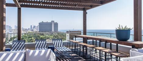 An outdoor deck with wooden furniture, a large potted plant, and a view of the city and ocean.