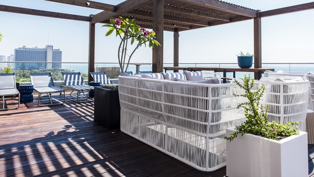 Een overdekt terras met witte rieten meubels en plantenbakken, met uitzicht op een stadslandschap en de zee.