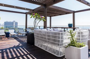Un espacio al aire libre cubierto con mobiliario de mimbre blanco y macetas, con vistas a la ciudad y al mar.