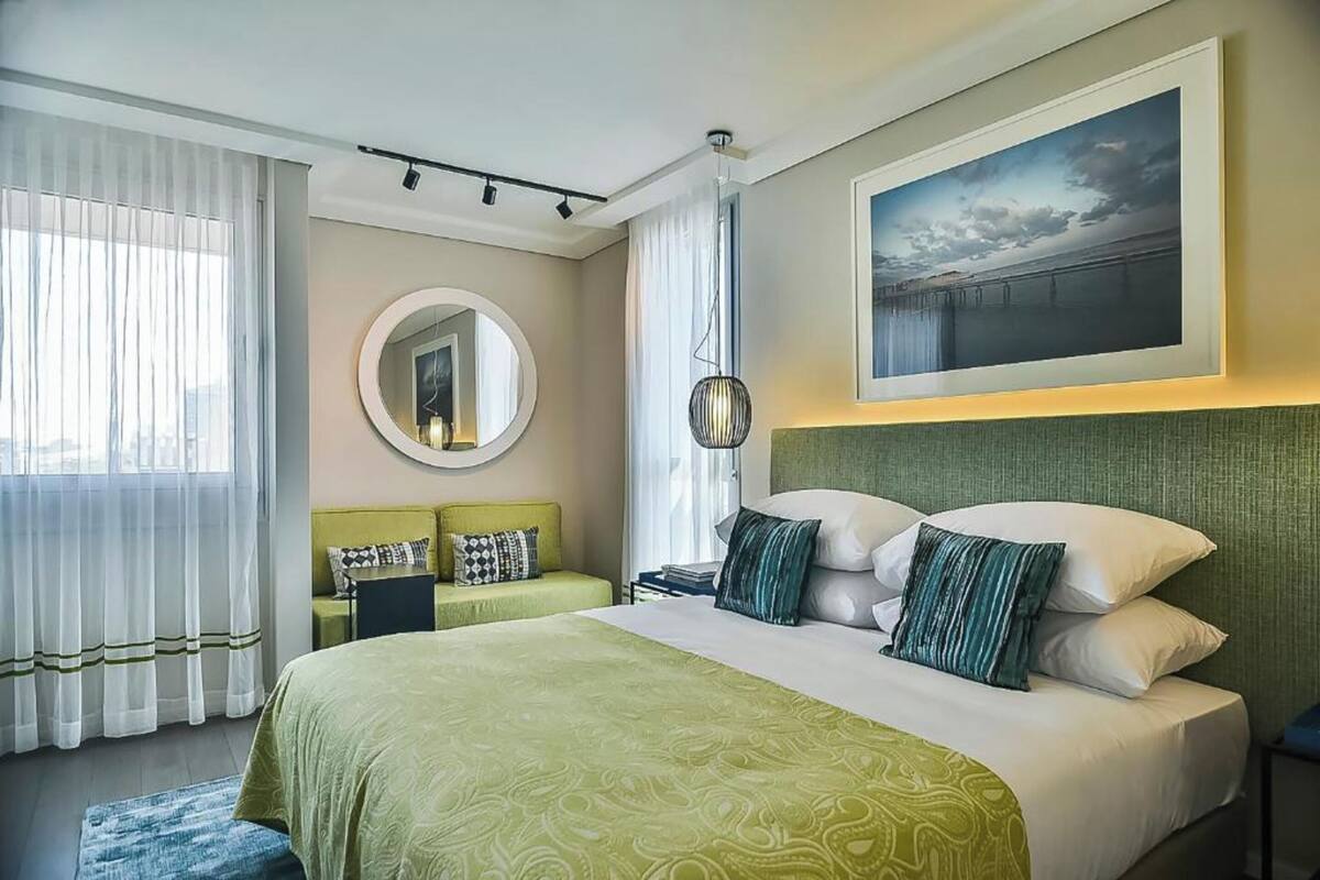 Une chambre d’hôtel avec un grand lit, un miroir rond, une tête de lit verte et un tableau accroché au mur.
