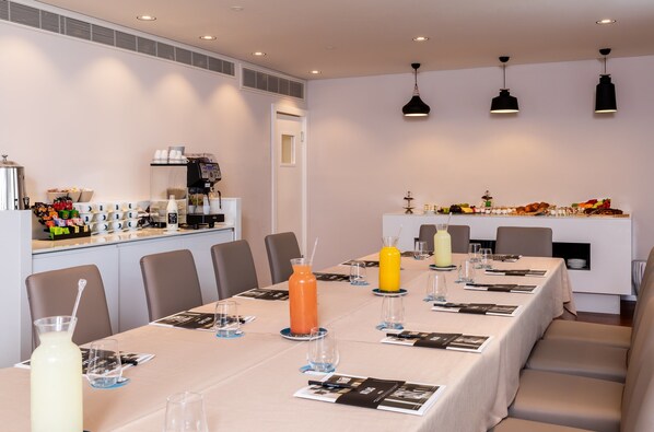Un salón de conferencias con una larga mesa dispuesta para una reunión, equipado con vasos de agua, una máquina de café y una zona de buffet con diferentes opciones de comida.