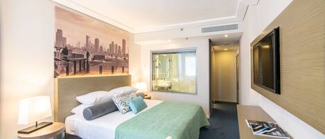 A hotel room with a bed, a desk, a chair, a TV, and a painting of a cityscape.