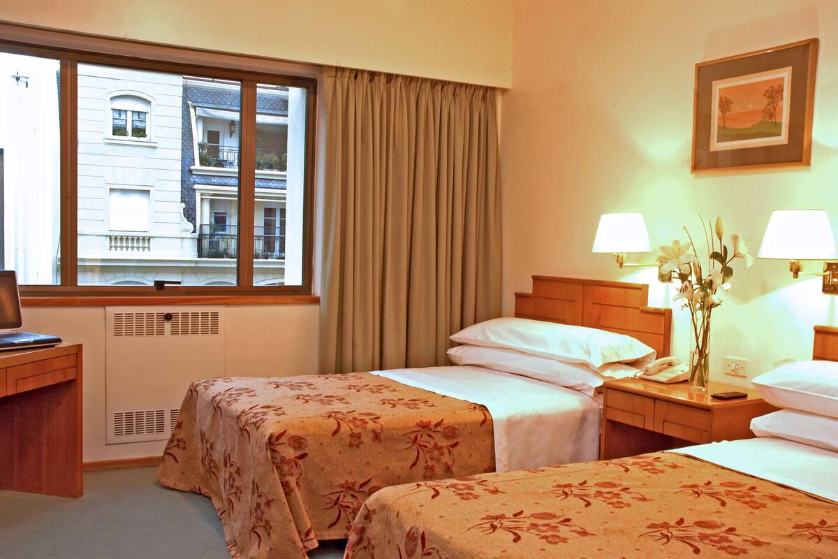 A hotel room with two beds, a desk, a window with curtains, and a picture on the wall.