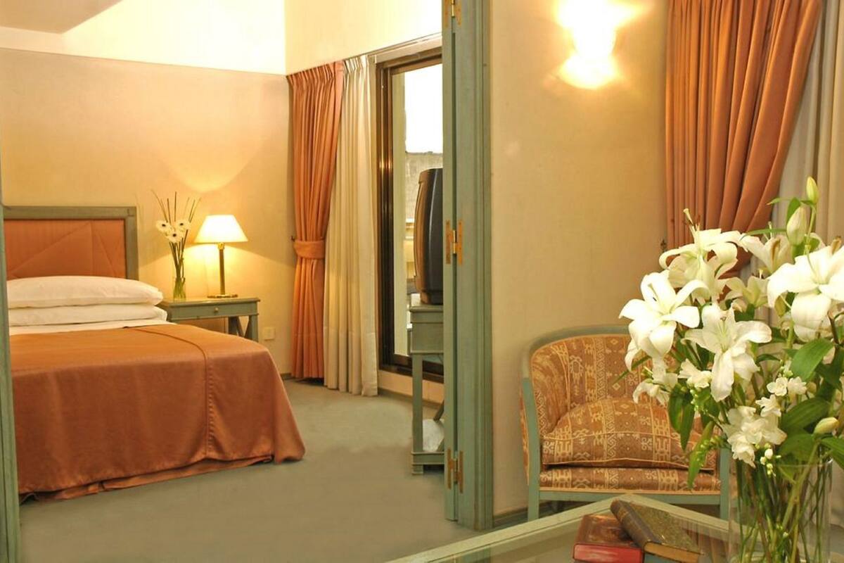 A hotel room with a bed, a nightstand with a lamp, a chair, and a mirror reflecting the room.