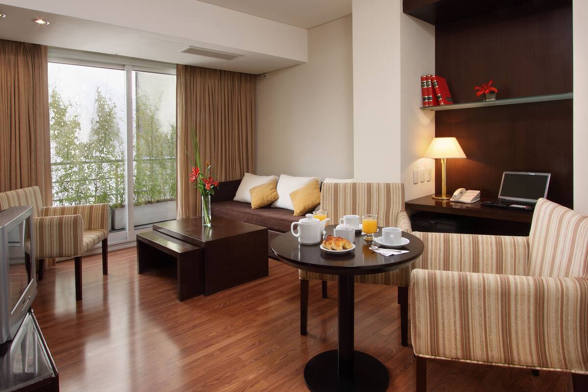 A modern hotel room with a sofa, armchairs, a dining table with breakfast, a TV, and a balcony with curtains.