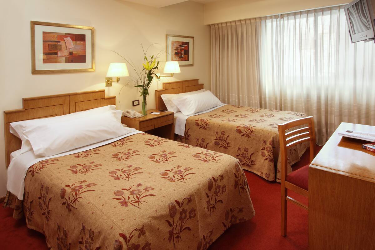 A hotel room with two beds, a desk, a chair, a lamp, and a window with curtains.