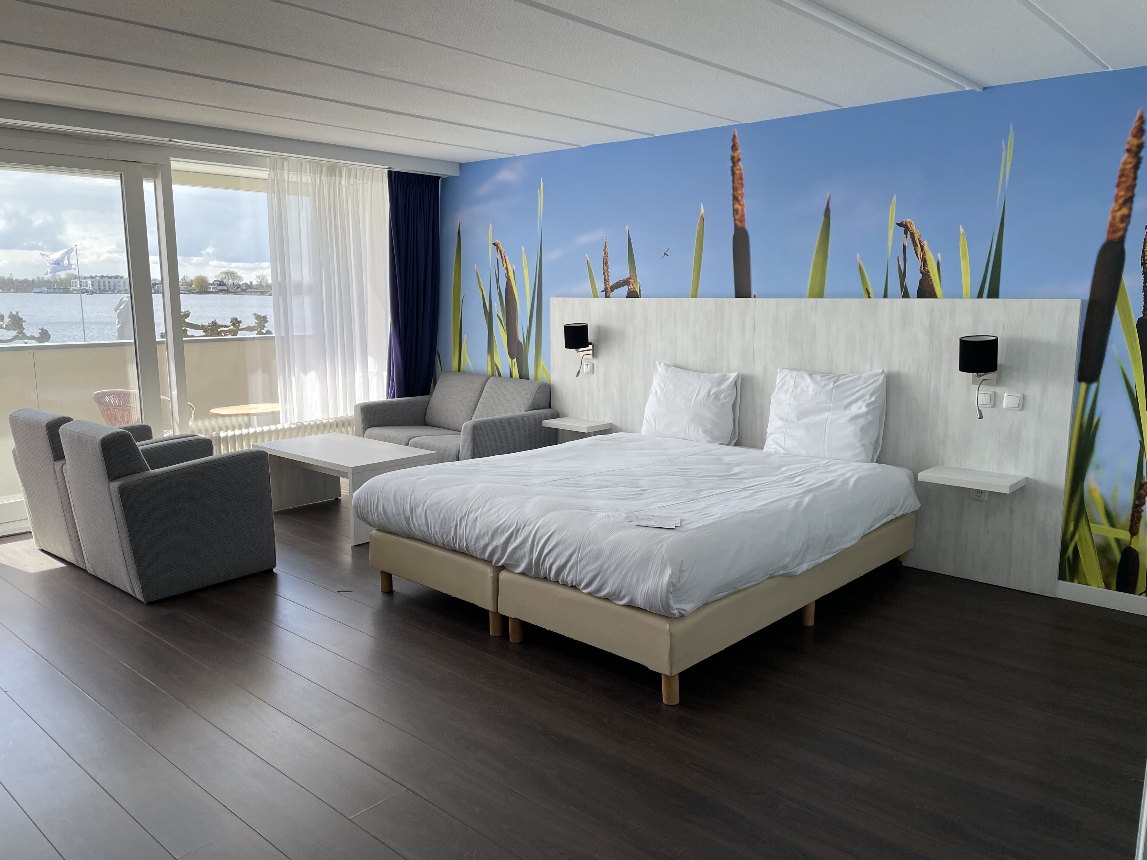 A modern hotel room with a large bed, a sofa, a small table, and a view of the sea.