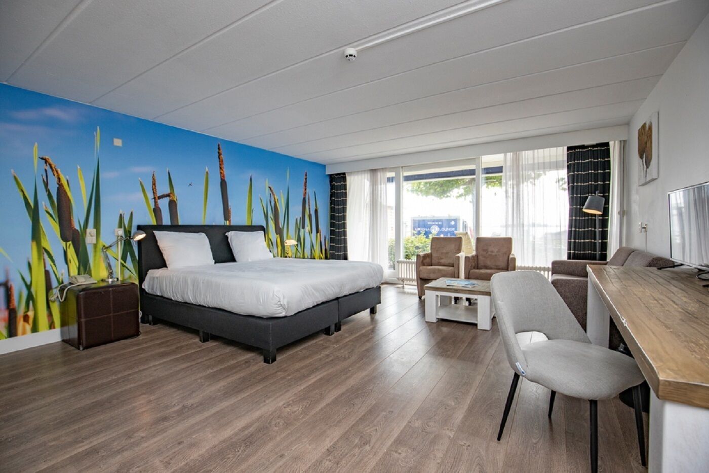 A modern hotel room with a bed, a desk, a chair, and a large window.