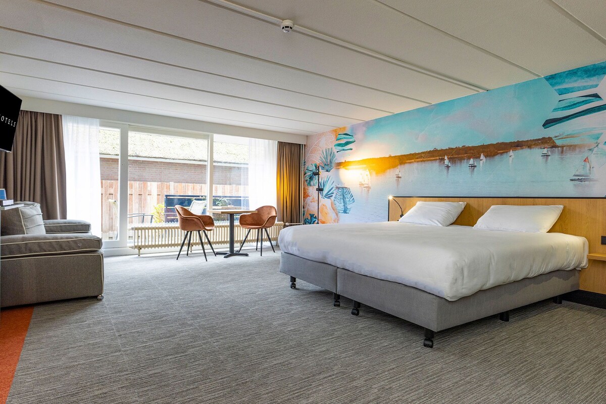 A hotel room with a bed, a sofa, two chairs, a large window, and a mural of a seascape.