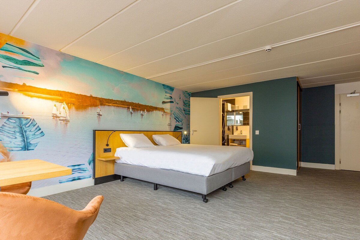 Une chambre d’hôtel comprenant un lit, un bureau, une chaise, une salle de bain avec un miroir et un mur orné d’un tableau.