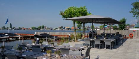 An outdoor patio with tables and chairs, a covered seating area, and a view of the water and buildings in the background.