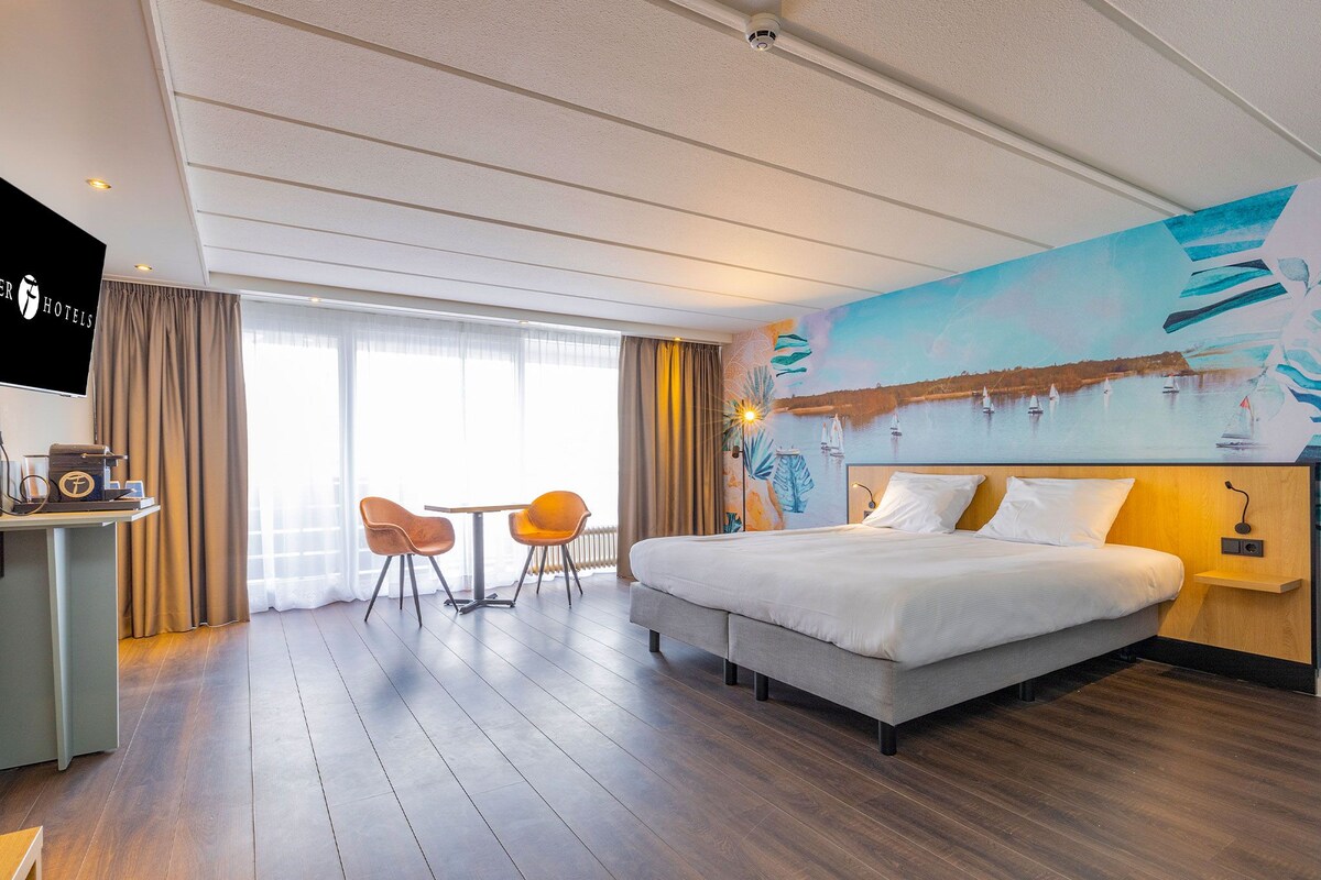 Une chambre d’hôtel comprenant un lit, un bureau, deux chaises, une télévision et une grande fenêtre avec des rideaux.