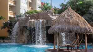 Zona de piscina con cascada, sillas de descanso y un pabellón con techo de paja.