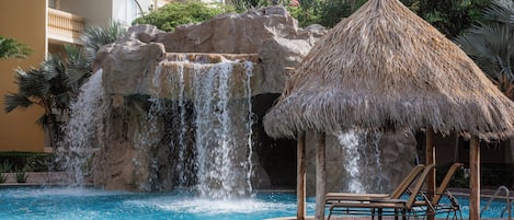 Zona de piscina con cascada, sillas de descanso y un pabellón con techo de paja.