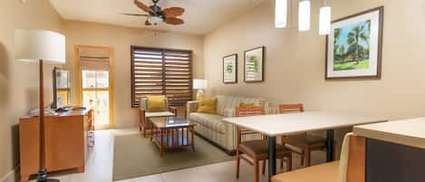 A well-lit living room with a sofa, dining table, and chairs. There is a ceiling fan, a lamp, and framed pictures on the wall.