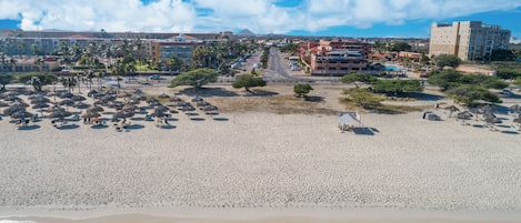 Une plage avec des parasols en chaume et des chaises longues, un hôtel à plusieurs étages doté de balcons, et une vue dégagée sur l’océan.