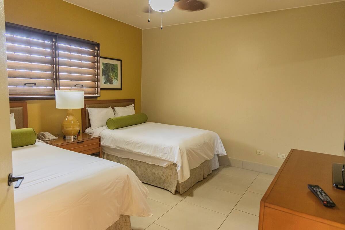 A hotel room with two beds, a nightstand with a lamp, a framed picture on the wall, and a remote control on a wooden table.