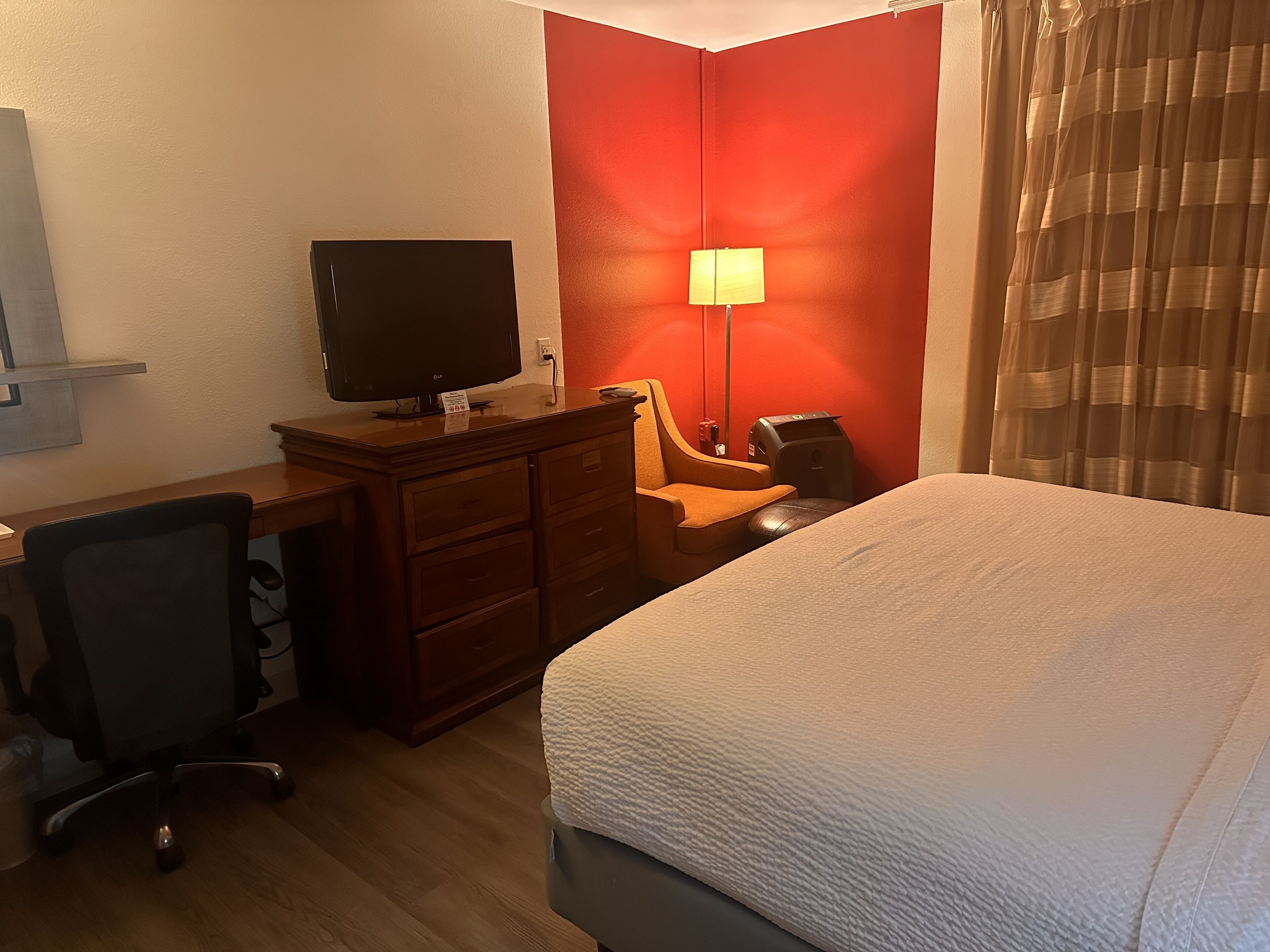 Camera d'albergo con un letto, una scrivania con televisore, una sedia, una lampada e una parete decorata in rosso.