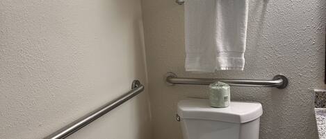 A bathroom with a toilet, a handrail, a towel rack, a trash bin, and a toilet paper holder.